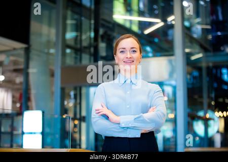 Selbstbewusste Geschäftsfrau mittleren Alters lächelt in der modernen Glasbürohalle in die Kamera, professionelles Firmenporträt mit natürlichem Licht. Stockfoto