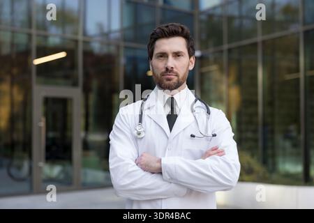 Junger erwachsener männlicher Arzt, der einen weißen Labormantel und ein Stethoskop mit überkreuzten Armen trägt und selbstbewusst vor einem modernen Krankenhaus- oder Klinikgebäude steht Stockfoto