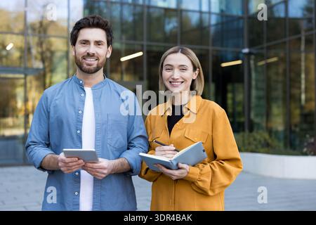 Verschiedene Geschäftspartner stehen vor einem modernen Bürogebäude, lächeln und halten ein Tablet und ein Notebook in der Hand, um Teamarbeit, Zusammenarbeit, Selbstvertrauen und Erfolg zu vermitteln Stockfoto