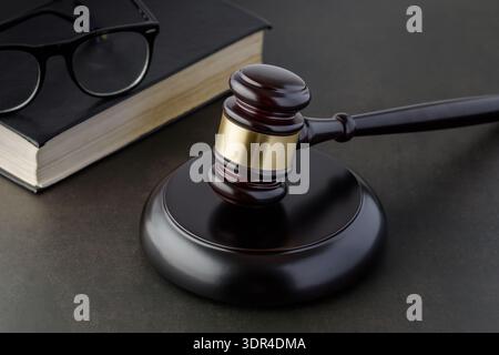 Brauner goldener hölzerner Hammer des Richters auf dunklem Tisch mit schwarzem Buch und Gläsern. Zubehör des Gerichts oder der Auktion. Stockfoto