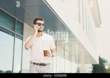Mann steht vor dem modernen Gebäude mit Kaffee, während er bei sonnigem Wetter telefoniert Stockfoto