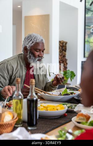 Afroamerikanische Familie trifft sich am Esstisch, Senior Mann inspiziert Salat mit Essstäbchen Stockfoto
