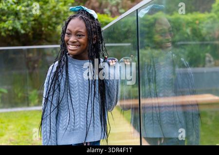 Afroamerikanisches Mädchen lächelt, hält Glasgeländer am Rasen, in Zopfstrick-Pullover und Schleife Stockfoto