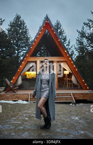 Die junge Frau genießt den Abend in der Nähe einer Hütte mit hellen Lichtern. Sie trägt ein Kleid und einen Mantel, während sie vor dem gemütlichen Haus lächelt, umgeben von Stockfoto