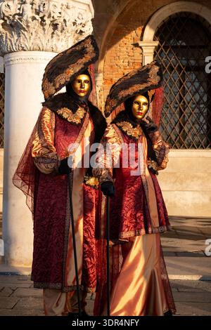 Paare in verzierten roten und goldenen Kostümen und Masken posieren vor dem Dogenpalast in San Marco während des jährlichen Karnevals in Venedig, Italien, für die Kamera. Stockfoto