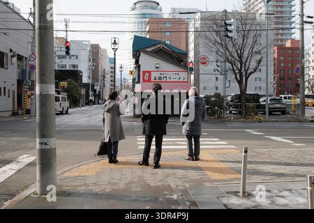 Drei Personen warten an einem Straßenübergang vor einem Ramen-Laden in Japan Stockfoto