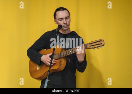 Ein junger Mann mit langen schwarzen Haaren steht vor einem gelben Hintergrund, spielt eine klassische akustische Gitarre und singt in ein Mikrofon. Stockfoto