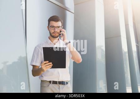 Geschäftsmann spricht tagsüber am Telefon, während er ein Tablet in der Nähe eines modernen Gebäudes hält Stockfoto