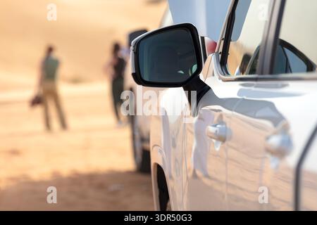 Regionale Abenteueridentität als Nahaufnahme des Seitenspiegels des SUV, der das Wüstenlicht in der Nähe von Dubai, VAE, reflektiert. Verschwommene Touristen im Hintergrund, Kontrast zu Mobili Stockfoto