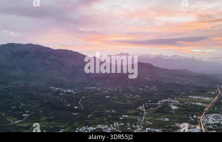 Panoramablick aus der Vogelperspektive Kretas Vorgebirge bei Sonnenuntergang mit schneebedeckten Gipfeln, Olivenhainen, Tal mit seeverstreuten Dörfern, gewundenen Straßen und Wolken Stockfoto