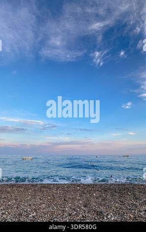 Kieselstrand und ruhiges Mittelmeer in Antalya, Türkei, mit kleinen Booten auf dem Wasser unter weichem, pastellfarbenen Abendhimmel. Stockfoto