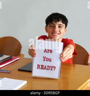 Sind Sie bereit? Zurück zur Schule. Ein junger Junge an seinem Schreibtisch, lächelt und zeigt ein Notizbuch vor die Kamera. Stockfoto