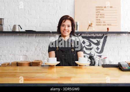 Barista serviert Kaffee in einem Café in gemütlicher Umgebung mit Holztheke und Menübrett Stockfoto