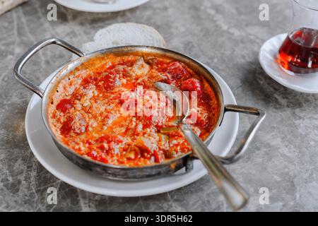 Shakshuka mit Eiern und Tomaten, serviert in einer heißen Metallpfanne auf einem Tisch mit Brot und Tee, fertig als warmes mediterranes Frühstück Stockfoto