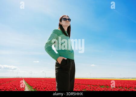Frau mit Sonnenbrille steht selbstbewusst über roten Tulpenreihen unter einem hellblauen Himmel und zeigt Frühlingsreisen, Lebensstil und Blumentourismus Stockfoto