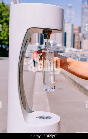 Die wiederverwendbare Flasche wird an einem Trinkbrunnen im Freien am Stadtaufer von Hand gefüllt und zeigt fließendes Wasser, nachhaltige Flüssigkeitszufuhr und alltägliche städtische Aktivitäten. Stockfoto