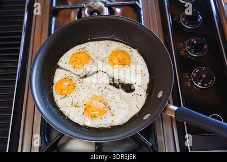 Vier sonnige Eier in einer schwarzen Pfanne auf einem heißen Herd anbraten, brutzeln, sprudeln und für ein einfaches hausgemachtes Frühstück kochen Stockfoto