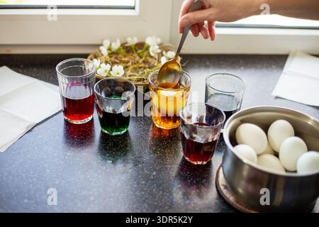 Eine Frau taucht mit einem Löffel ein weißes Ei in ein Glas Orangenfarbe auf einer Küchenzeile. osterzubereitung, Färben von Eiern, Feiertagstradition. Ostern. Stockfoto
