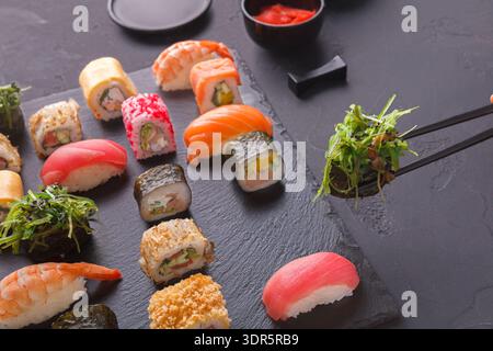 Sushi-Collage auf einer schwarzen Steinplatte mit Stäbchen, die Garnelen aufnehmen Stockfoto