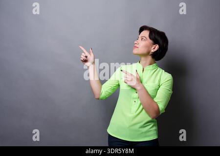 Frau mit grünem Shirt zeigt nach rechts vor grauem Hintergrund in einem Studio-Setting Stockfoto