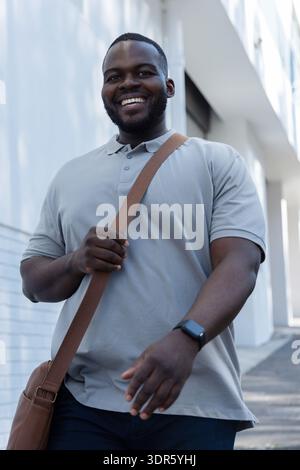Afroamerikaner, der auf einem Weg an der weißen Fassade zur Kamera läuft, einen Taschenriemen hält, eine Uhr trägt Stockfoto