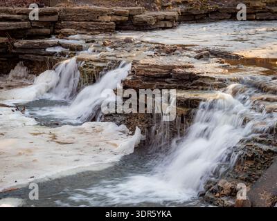 Wasserfall, der über Kalksteinschichten mit schmelzendem Eis während des Frühjahrs unter warmem Sonnenlicht fließt Stockfoto