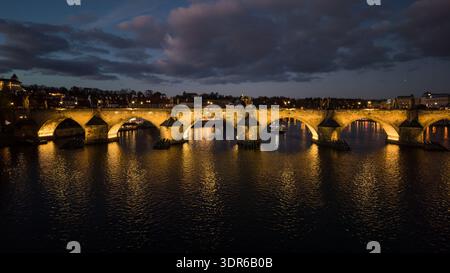 Die Luftaufnahme zeigt die Karlsbrücke in Prag bei Nacht beleuchtet. Die Brücke überspannt den Fluss mit Booten auf dem Wasser und Gebäuden entlang des Ufers. Stockfoto