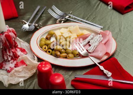 Nahaufnahme eines Vorspeisetellers mit grünen Oliven, Käsewürfeln, geräucherten Salamistöcken und gefaltetem Schinken, auf einem rustikalen weißen Teller mit Re angeordnet Stockfoto