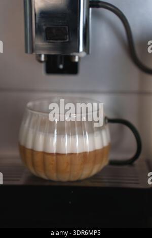 Nahaufnahme eines Glasbechers mit mehrschichtigem Kaffee und Schaum auf einer Espressomaschine in moderner Küche, die die cremige Textur und handwerkliche Verarbeitung unterstreicht Stockfoto