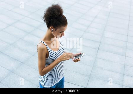Die junge Frau hört Musik auf ihrem Telefon, während sie auf einer gekachelten Fläche in einem Stadtgebiet steht Stockfoto