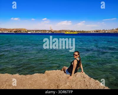 Europäischer Tourist sitzt am Rande einer Klippe über dem türkisblau-azurblauen Wasser des Roten Meeres in Scharm El Sheikh (Ägypten). Eine junge Frau in Sommerkleidung Stockfoto