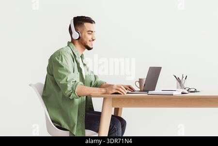 Der junge Mann arbeitet am Schreibtisch, während er tagsüber Kopfhörer in hellen Räumen trägt Stockfoto