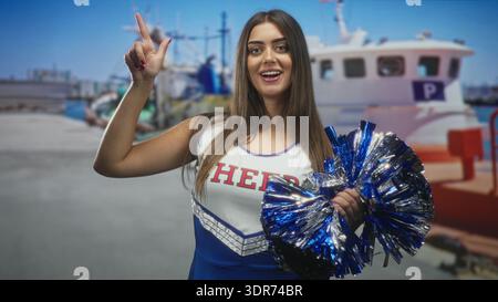 Cheerleaderin, die im Studio glänzende blaue Pompom-Punkte mit dem Zeigefinger nach oben hält; aufregender Teamgeist. Stockfoto
