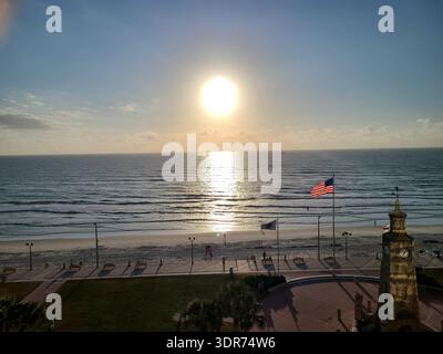 NUR REDAKTIONELLE VERWENDUNG - DAYTONA BEACH FLORIDA: ​A atemberaubende Aussicht auf die Atlantikküste am Daytona Beach, Florida, mit dem berühmten Coquina Clock Tower. Die Sonne hängt tief und hell an einem klaren blauen Himmel und erzeugt eine brillante goldene Reflexion auf der Meeresoberfläche. Eine amerikanische Flagge weht im Vordergrund in der Meeresbrise und umrahmt die friedliche Szene des Strandes, der Promenade und des historischen Steinmauerwerks des Uhrenturms während der goldenen Stunde. Stockfoto