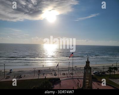 NUR FÜR REDAKTIONELLE ZWECKE - DAYTONA BEACH FLORIDA: ​A aus einem Hochwinkel mit malerischem Blick auf den historischen Coquina Clock Tower am Daytona Beach Boardwalk in Florida, USA. Die helle Morgensonne liegt in einem trüben, wolkenbedeckten Himmel, der eine glänzende Reflexion über die sanften Wellen des Atlantischen Ozeans wirft. Im Vordergrund weht eine amerikanische Flagge in der Küstenbrise neben dem Steinturm und überblickt einen weiten weißen Sandstrand und eine ruhige Fußgängerpromenade, während der Tag beginnt. Stockfoto