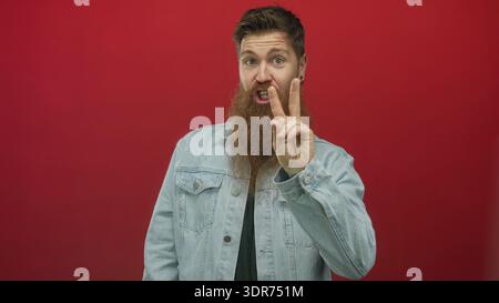Mann mit langem Bart, der Denimjacke über schwarzem Hemd trägt, winkt Hand im roten Studio und blickt nach oben; Glück. Stockfoto