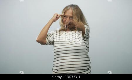 Die ältere blonde Frau zeigt den Finger direkt auf die Kamera, während sie Gesten auf ihren Tempel drückt, und trägt ein gestreiftes Hemd im Studio; eine selbstbewusste Herausforderung. Stockfoto