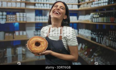Bäckerin hält einen runden Kuchen mit Händen, lächelt und lacht, während sie eine Schürze in einem Ladengebäude trägt; Freude Gastfreundschaft Wärme. Stockfoto