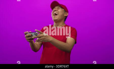 Junger Mann mit roter Mütze und T-Shirt, der das Smartphone hält und lacht, während er ein mobiles Spiel im magentafarbenen Studio spielt; Spaß am Spaß. Stockfoto