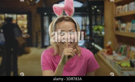 Frau in rosa Hemd mit Hasenohren lächelt und hält den Finger an die Lippen, um im Gebäude Ruhe zu finden; ruhige Geheimhaltung. Stockfoto