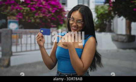 Junge Brünette, die eine Kreditkarte hält und auf der Straße mit dem Finger auf die Karte zeigt, die neben Geländer und Bougainvillea blau oben trägt; Selbstvertrauen Stockfoto