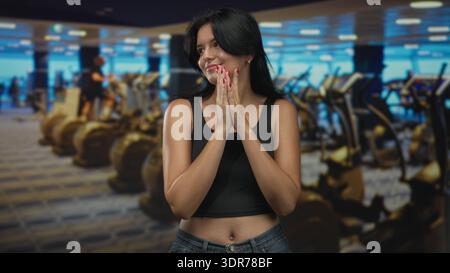 Eine Frau mit gefalteten Händen, die schwarze Crop-Top trägt, lächelt im Fitnessstudio zwischen Laufbändern und Ellipsentrainer hell; Gelassenheit. Stockfoto