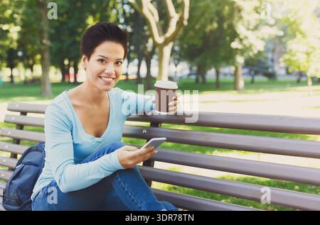 Frau, die tagsüber auf einer Bank im Park sitzt und Kaffee genießt und ein Smartphone benutzt Stockfoto