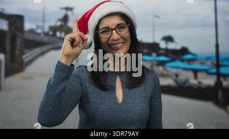 Frau mit weihnachtsmütze und -Brille zeigt den Finger nach oben auf den Strandweg, an den Sonnenschirmen am Meer; Feiertagsstimmung. Stockfoto