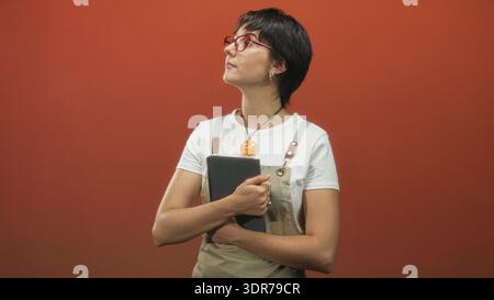 Eine Frau mit Schürze und roter Brille hält eine schwarze Tafel an der Brust, die Arme um sie gewickelt, ein orangener Anhänger sichtbar an einer orangen Studiowand; t Stockfoto