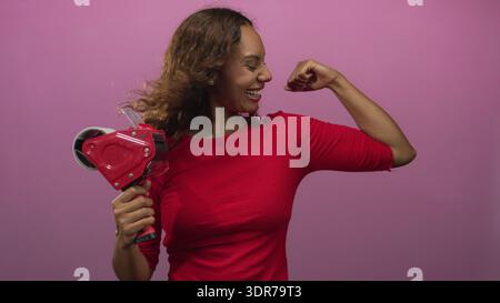 Frau mit Klebebandspender und flexiblem Bizeps im Studio mit rosa Hintergrund und breitem lachenden Lächeln; Selbstvertrauen-Feier. Stockfoto