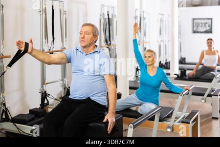 Fokussierter Mann und zwei Frauen unterschiedlichen Alters, die Dehnungsübungen machen, während sie Pilates-Bettgerät im Rehabilitationszentrum benutzen Stockfoto