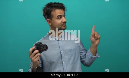 Junger hispanischer Mann mit Kamera, der auf etwas auf isoliertem grünen Hintergrund zeigt, was auf Idee oder Richtung hindeutet Stockfoto