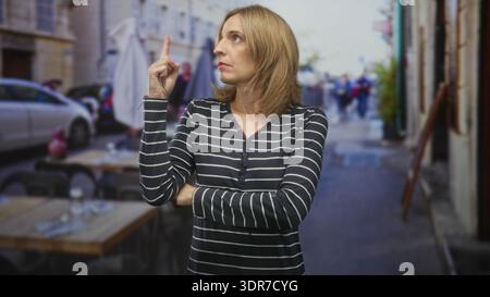 Frau zeigt mit gefalteten Armen den Finger nach oben auf den Straßencafé-Tisch und schaut zur Seite; Ungeduld. Stockfoto