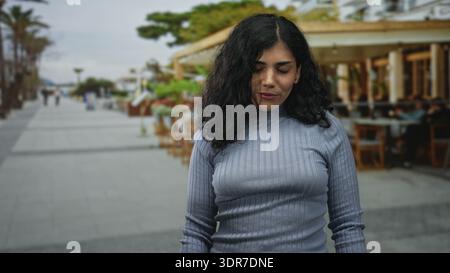 Junge brünette Frau, die mit geschlossenen Augen und einem leichten Lächeln auf der Straße neben einer Restaurantterrasse im Freien steht; ruhige Reflexion. Stockfoto